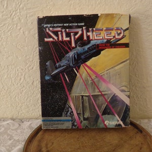 Op de afbeelding: Een vintage gamebox voor het spel Silpheed, met een ruimteschip dat door een futuristisch stadslandschap vliegt. De doos is zwart met rode en witte accenten en heeft de tekst "JAPAN'S HOTTEST NEW ACTION GAME" en "FROM THE DESIGNERS OF THEXDER" erop.