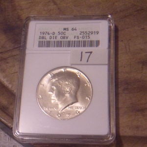 May include: A silver 1974-D 50C half dollar coin graded MS 64 in a plastic coin holder with a label that reads "DBL DIE OBV FS-015" and "2552919".