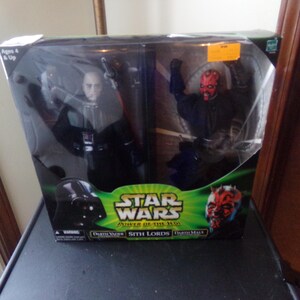 Op de afbeelding: Een Star Wars Power of the Force Sith Lords action figure set met Darth Vader en Darth Maul. De figuren zijn verpakt in een groene, zwarte en rode doos met het Star Wars-logo.