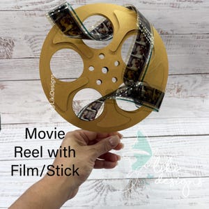 以下が含まれることがあります： 金色の映画リールにフィルムストリップが付いています。リールは円形のデザインで、円がくり抜かれており、透明な棒で支えられています。フィルムストリップは白黒です。「Movie Reel with Film/Stick」というテキストが表示されています。