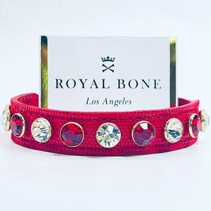May include: A red dog collar with a textured pattern, decorated with alternating red and clear gemstones set in gold-coloured bezels. The collar features the text "ROYAL BONE Los Angeles" on a white card.
