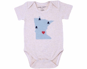 May include: A beige baby onesie with a blue outline of the state of Minnesota with three pine trees and a red heart in the center. The onesie has the text "Baby Buffalo" and "3-6m" printed on the front.