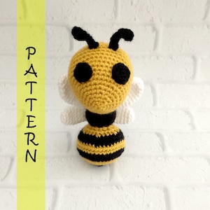 May include: A crocheted bee toy with black and yellow stripes, white wings, and black eyes. The bee is standing on a white brick background.