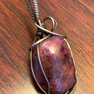 May include: A silver wire wrapped pendant featuring a pink and purple gemstone with a speckled pattern.