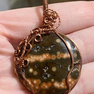 May include: A handcrafted pendant featuring a polished, round gemstone with a speckled pattern in shades of green, brown, and cream. The stone is encased in intricate copper wire, forming a decorative frame and bail for hanging.