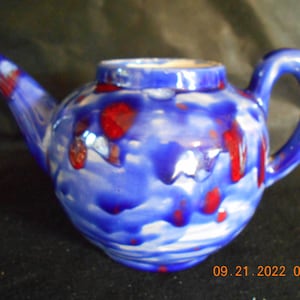 May include: A blue and red ceramic teapot with a white glaze. The teapot has a round body and a curved spout. The handle is on the side of the teapot.