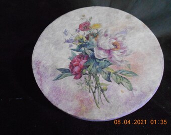 May include: A round, white, paper-based coaster with a floral design. The design features pink, white, and yellow flowers with green leaves. The coaster has a textured surface and a light purple background.