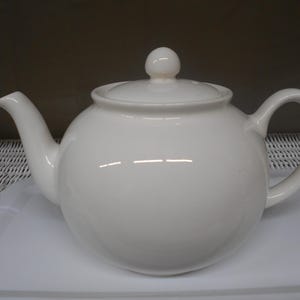 May include: A white ceramic teapot with a rounded body, a curved spout, and a loop handle. The lid has a small, round knob. The teapot sits on a white tray. The teapot is a classic design.
