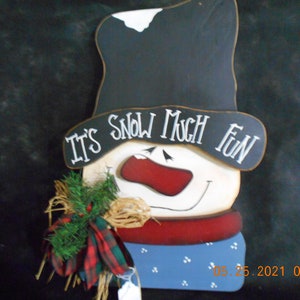 May include: A wooden snowman decoration with a black top hat and a white face. The snowman has a red nose and a red and blue scarf. The text "It's Snow Much Fun" is written on the hat.