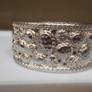 May include: A silver-colored cuff bracelet with a textured surface. The bracelet features a repeating pattern of raised, circular designs and a rope-like border. The jewelry is made of a shiny, metallic material.