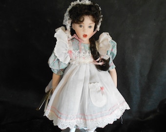 May include: A porcelain doll with long dark hair styled in a braid, wearing a white dress with a blue and pink floral print, a white apron, and a white lace bonnet.
