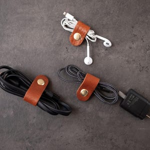 May include: Three tan leather cable organizers with gold-toned metal snap buttons. Each organizer is wrapped around a different type of electronic cord, including a white pair of earbuds, a thick black cable, and a black and white braided cord.