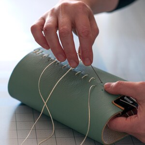 A person's hands are carefully sewing a green leather item, possibly a wallet or a bag, using white thread.