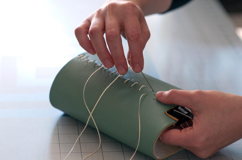 A person's hands are carefully sewing a green leather item, possibly a wallet or a bag, using white thread.