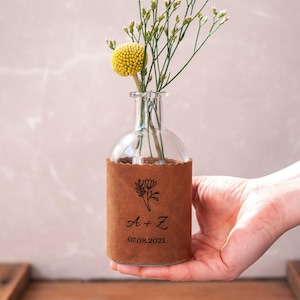 May include: A clear glass vase with a brown paper sleeve, holding a yellow flower and sprigs of greenery. The paper sleeve is printed with a floral design, initials, and the date 07.08.2021. The vase is held in a hand.