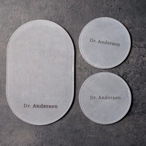 May include: A set of three gray leather coasters. One oval and two round coasters are visible. Each coaster has a stitched border and the name "Dr. Anderson" printed on it. The coasters are arranged on a dark gray surface.