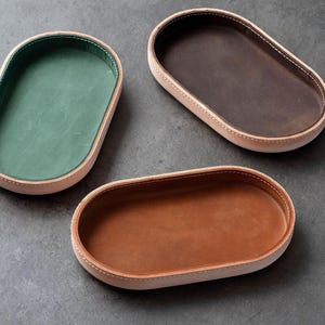 May include: Three oval-shaped valet trays with light tan leather outer walls and contrasting interior linings in forest green, dark brown, and medium tan. Each tray features visible white stitching along the rim and a smooth, matte finish.