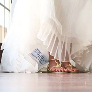 Può includere: Un abito da sposa in tulle bianco con gonna a strati e una piccola toppa in tessuto bianco cucita sull'orlo interno. La toppa presenta un testo nero scritto a mano: "date: 6/25/16, name: kyle & ashley, wedding date: 6/25/16, love you always, mom & dad".