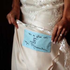 Può includere: Abito da sposa color crema con dettagli in pizzo e una toppa azzurra cucita all'interno. La toppa presenta un testo scritto a mano con inchiostro nero che recita "I'm so glad it's you xoxo Mom Chiaramonte."
