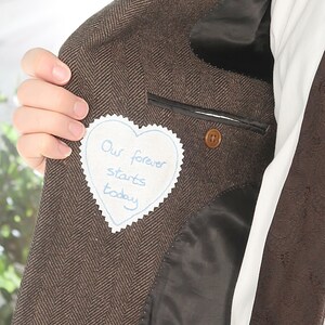Può includere: Una giacca di tweed marrone con una toppa a forma di cuore cucita sulla tasca interna. La toppa è bianca con cuciture blu e dice "Il nostro per sempre inizia oggi".
