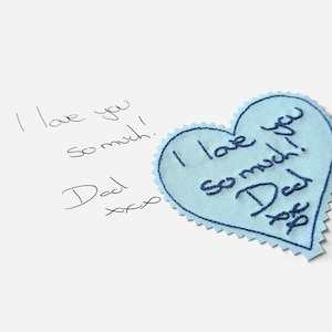 Könnte beinhalten: Ein hellblauer herzförmiger Stoff-Patch mit dem Text "I love you so much! Dad xoxo", der in dunkelblauem Garn gestickt ist.