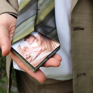 Könnte beinhalten: Eine grün-schwarze gestreifte Krawatte mit einem Foto, das in die Falte gesteckt ist. Das Foto zeigt eine Frau, die ein Baby hält.