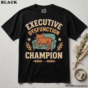Flat lay of a black Comfort Colors 1717 garment-dyed neurodivergent cat shirt on a stone background with dried florals. A vintage 1970s graphic depicts a lazy orange cat resting on an armchair with bold text reading "Executive Dysfunction Champion".