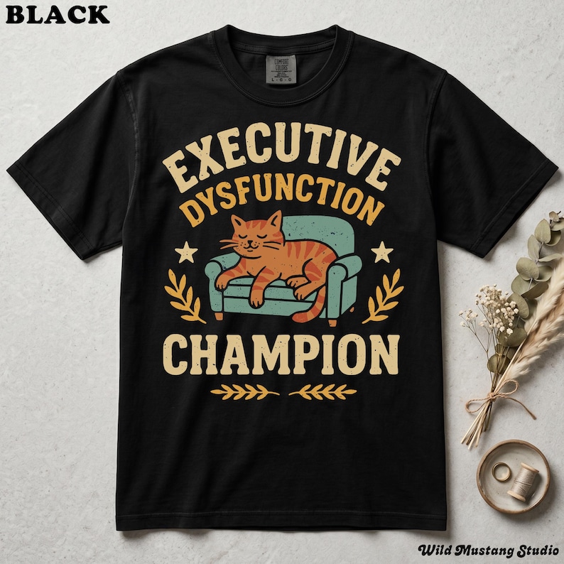 Flat lay of a black Comfort Colors 1717 garment-dyed neurodivergent cat shirt on a stone background with dried florals. A vintage 1970s graphic depicts a lazy orange cat resting on an armchair with bold text reading "Executive Dysfunction Champion".