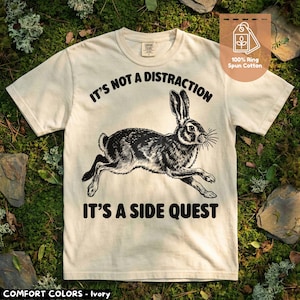 Puede incluir: Camiseta de color marfil con un gráfico negro de una liebre corriendo y el texto "IT'S NOT A DISTRACTION IT'S A SIDE QUEST." La camiseta está hecha de 100% algodón hilado en anillo. La marca "COMFORT COLORS" está impresa en la parte inferior.