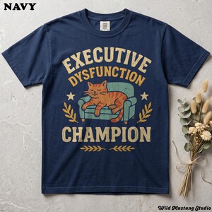 Flat lay of a Navy Comfort Colors 1717 garment-dyed neurodivergent cat shirt on a stone background with dried florals. A vintage 1970s graphic depicts a lazy orange cat resting on an armchair with bold text reading "Executive Dysfunction Champion".