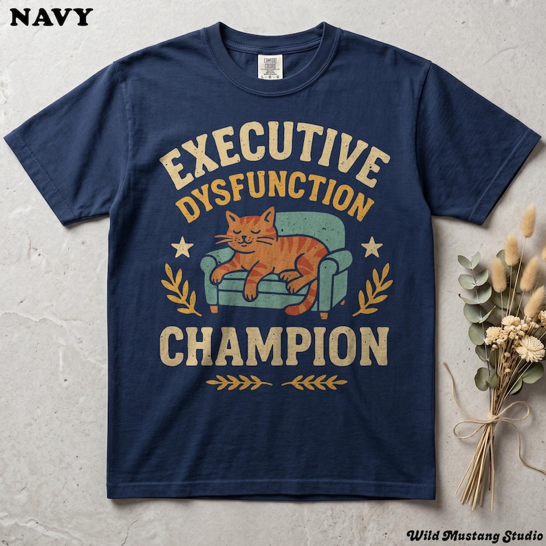 Flat lay of a Navy Comfort Colors 1717 garment-dyed neurodivergent cat shirt on a stone background with dried florals. A vintage 1970s graphic depicts a lazy orange cat resting on an armchair with bold text reading "Executive Dysfunction Champion".