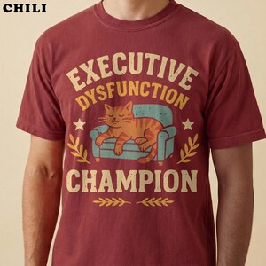 Closeup of a model wearing a chili red executive dysfunction shirt. The distressed retro graphic shows an orange tabby cat sleeping on a teal couch with funny text reading "Executive Dysfunction Champion" in cream and gold lettering.