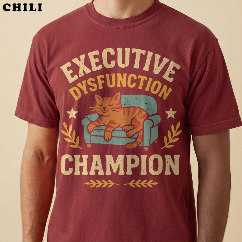 Closeup of a model wearing a chili red executive dysfunction shirt. The distressed retro graphic shows an orange tabby cat sleeping on a teal couch with funny text reading "Executive Dysfunction Champion" in cream and gold lettering.