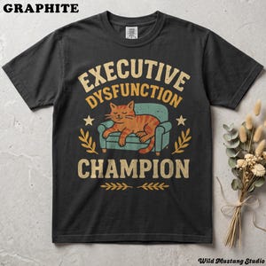 Flat lay of a Graphite or dark grey garment-dyed neurodivergent cat shirt on a stone background with dried florals. A vintage 1970s graphic depicts a lazy orange cat resting on an armchair with bold text reading "Executive Dysfunction Champion".
