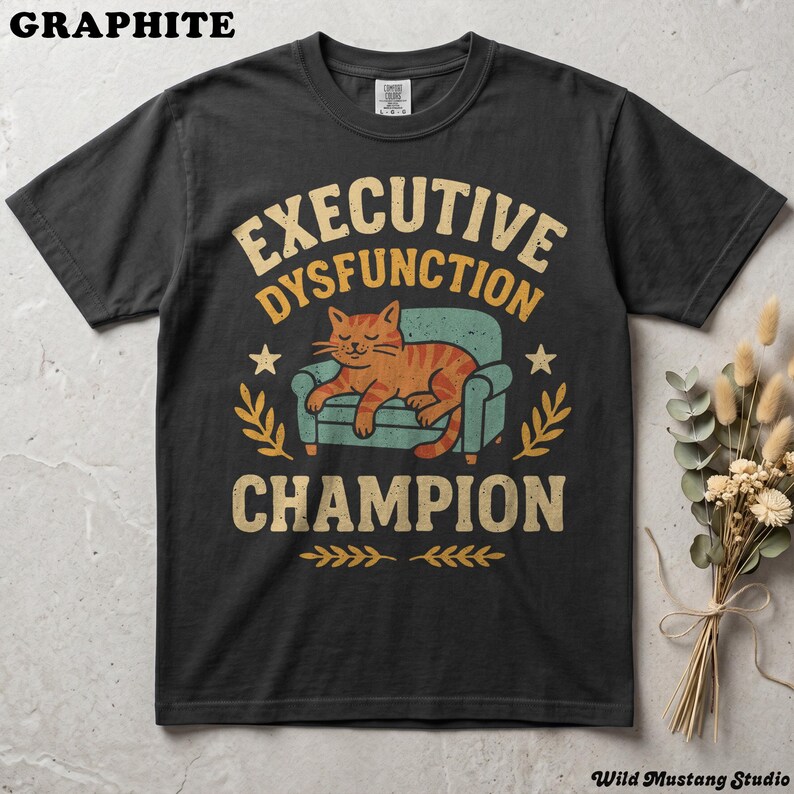 Flat lay of a Graphite or dark grey garment-dyed neurodivergent cat shirt on a stone background with dried florals. A vintage 1970s graphic depicts a lazy orange cat resting on an armchair with bold text reading "Executive Dysfunction Champion".