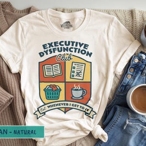 Può includere: T-shirt color crema con la scritta "Executive Dysfunction Club". Il design include un libro, una lista di cose da fare, un cesto per la biancheria e una tazza di caffè. La maglietta è abbinata a jeans blu e una tazza di caffè.