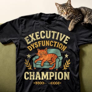 Peut inclure: T-shirt noir avec le texte "EXECUTIVE DYSFUNCTION CHAMPION" dans une police vieillie. Le graphisme représente un chat de dessin animé se prélassant sur un petit canapé. Un chat tigré repose sur le t-shirt.