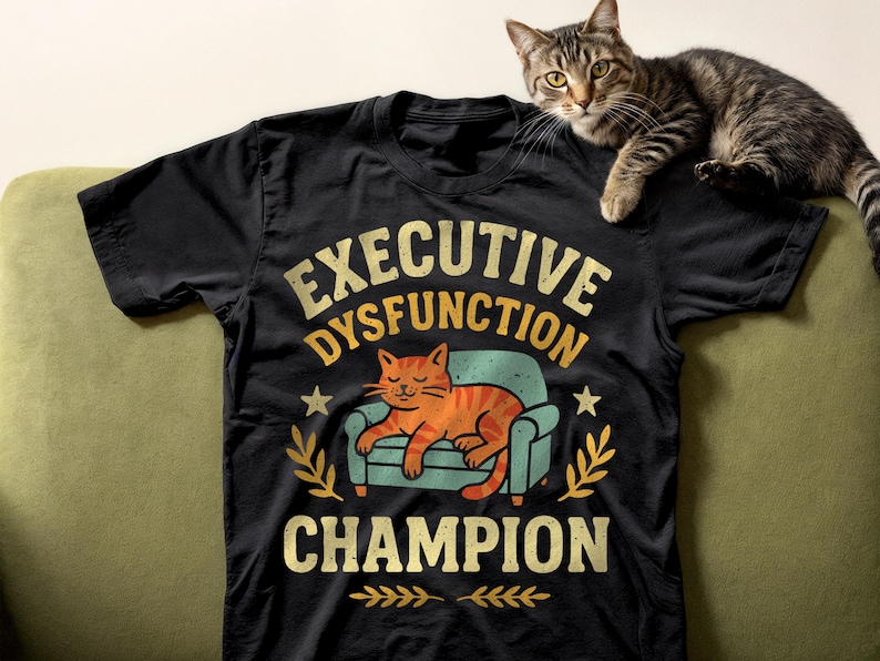 Black comfort colors style t-shirt with the text "EXECUTIVE DYSFUNCTION CHAMPION" in a distressed font. The graphic features a cartoon cat lounging on a small couch. A tabby cat rests on the shirt in the thumbnail