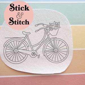 May include: A black and white line drawing of a bicycle with a basket of flowers. The bicycle is on a white, circular patch with the words "Stick & Stitch" in a pink circle.