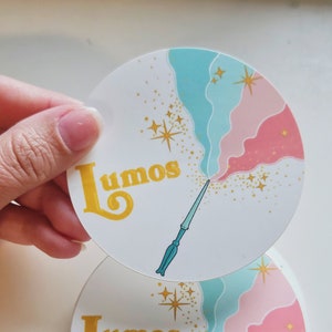 Puede incluir: Dos pegatinas redondas con fondo blanco, remolinos azules y rosas y purpurina dorada. Las pegatinas presentan una varita azul con mango dorado y la palabra "Lumos" en letras doradas.