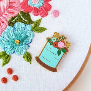 May include: A white embroidery hoop with a floral design embroidered in pink, yellow, green, and blue thread. A gold-tone enamel pin of a spool of thread with flowers on top is attached to the hoop.
