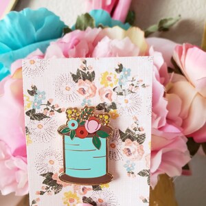 May include: A gold-tone enamel pin featuring a spool of thread with a floral arrangement on top. The spool is blue with a brown base. The flowers are pink, red, yellow, and blue.