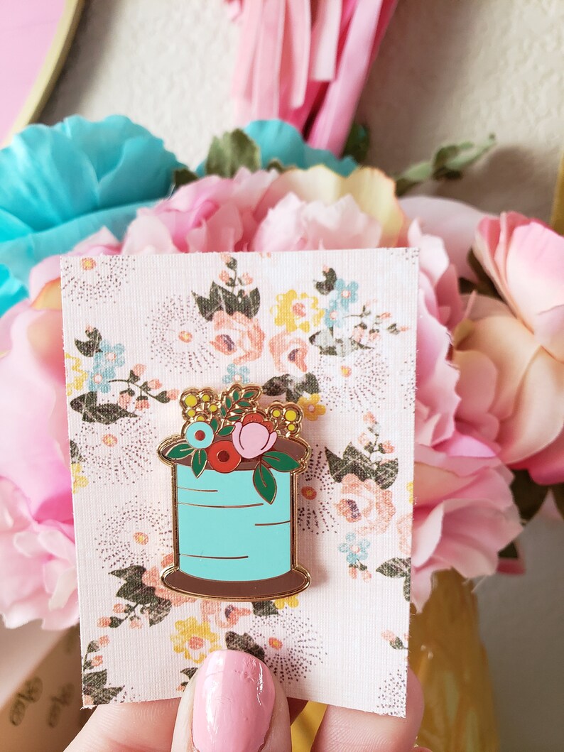May include: A gold-tone enamel pin featuring a spool of thread with a floral arrangement on top. The spool is blue with a brown base. The flowers are pink, red, yellow, and blue.