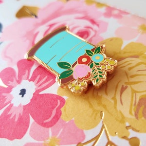 May include: A gold-tone enamel pin featuring a spool of thread with a floral design. The spool is light blue with a brown base and is adorned with pink, red, yellow, and blue flowers.