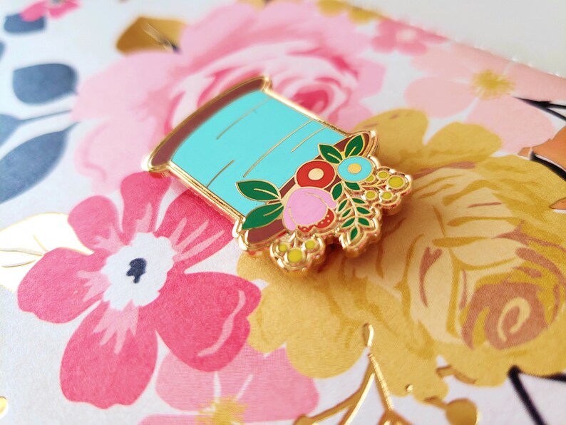 May include: A gold-tone enamel pin featuring a spool of thread with a floral design. The spool is light blue with a brown base and is adorned with pink, red, yellow, and blue flowers.