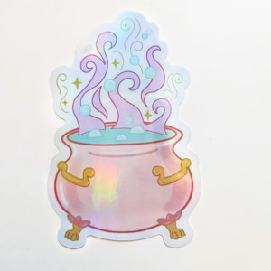 Puede incluir: Un caldero rosa con detalles dorados y humo morado y azul que sale de la parte superior. El caldero está sobre tres patas y tiene un líquido azul claro en su interior.
