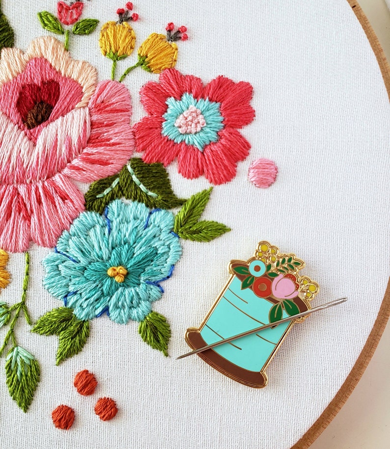May include: A gold-toned enamel pin shaped like a spool of thread with a needle. The spool is turquoise blue and has a floral design with pink, red, and yellow flowers. The needle is silver and is threaded through the spool.