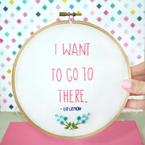 Puede incluir: Arte de bordado en aro con el texto "I want to go to there. - Liz Lemon" en hilo rosa. El aro está sobre un fondo blanco y una superficie rosa claro.