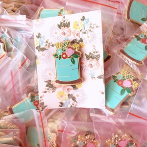 May include: A collection of gold-tone enamel pins featuring a spool of thread with flowers. The spools are turquoise blue with gold accents and the flowers are pink, red, and yellow.