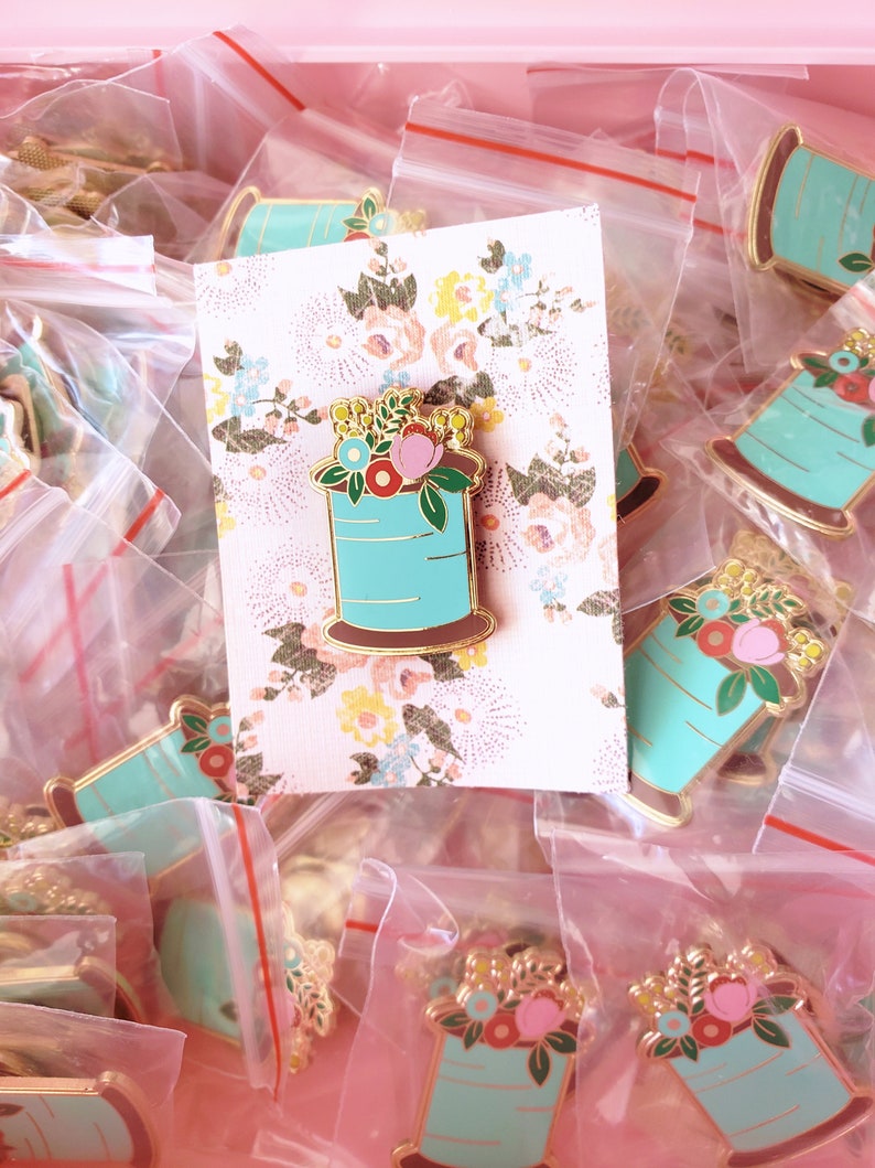 May include: A collection of gold-tone enamel pins featuring a spool of thread with flowers. The spools are turquoise blue with gold accents and the flowers are pink, red, and yellow.